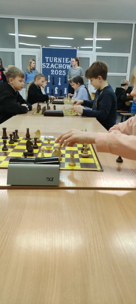 na zdjęciu widać grupa kilku chłopców siedzi przy stolikach na których są rozłożone plansze do gry w szachy. Na planszach stoją figury szachowe , obok zegary czarne mierzące czas gry uczestników turnieju szachowego. W oddali widać dwie panie stojące obok tablicy z napisem Turniej Szachowy 2025