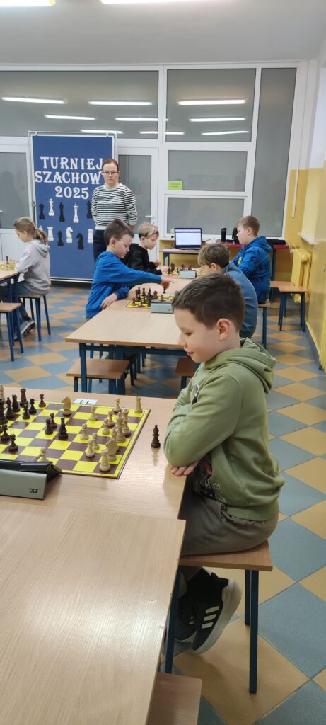na zdjęciu widać kilku chłopców siedzących przy stolikach, na których leżą plansze szachowe i figiry szachowe. Na pierwszym planie widać chłopca w zielonej bluzie grającego w szachy. W oddali widać stojącą tablicę z napisem Turniej Szachowy a obok stoi pani.