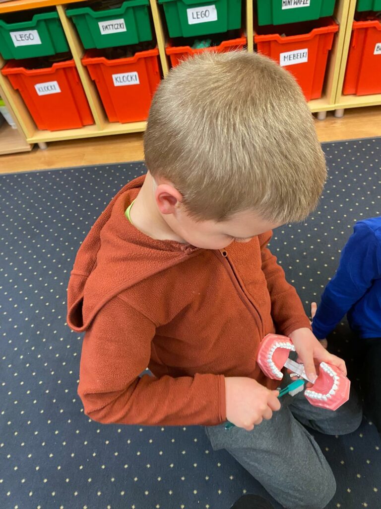 zdjęcie przedstawia chłopca w pomarańczowej bluzie, który siedzi na granatowym w białe kropki dywanie. W ręce lewej trzyma model jamy ustnej, a w prawej szczoteczkę do zębów, którą szczotkuje zęby górnego podniebienia w modelu. W tle widać pudełka zielone i czerwone z zawartością opisaną napisami na białym tle