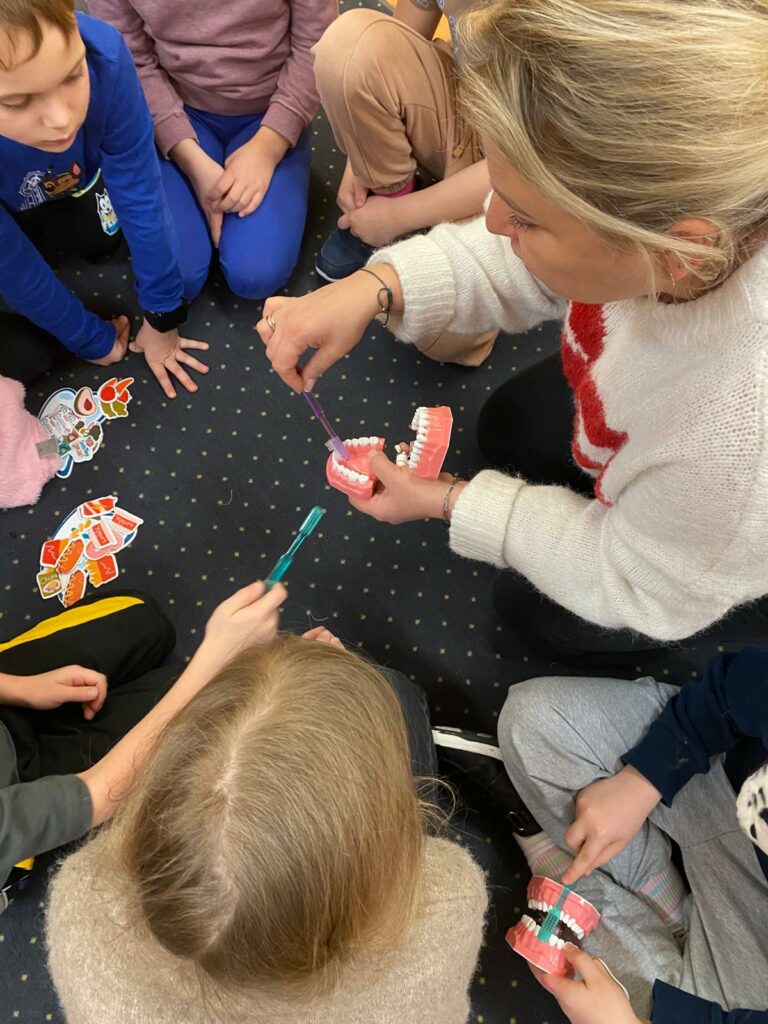na zdjęciu pani higienistka w blond włosach w białym sweterku siedzi na dywanie i trzyma w prawej ręce szczoteczkę do zębów, a w lewej ręce model jamy ustnej. Pokazuje siedzącym na dywanie dzieciom prawidłowe mycie zębów na modelu.