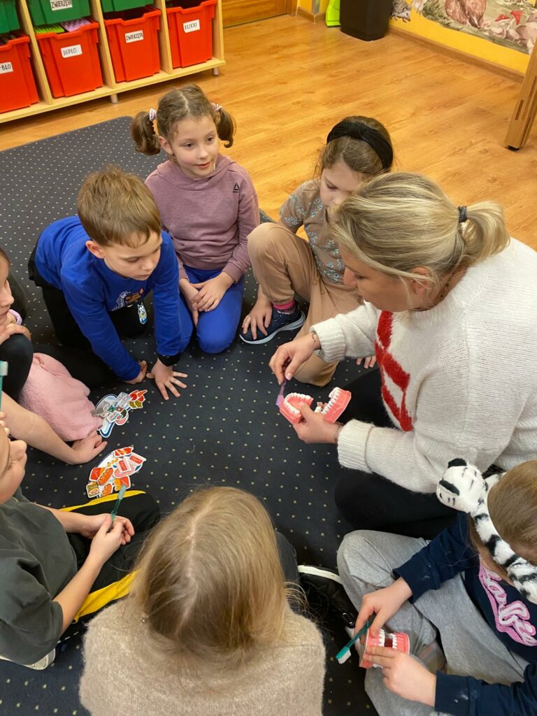 na zdjęciu pani higienistka w blond włosach w białym sweterku siedzi na dywanie i trzyma w prawej ręce szczoteczkę do zębów, a w lewej ręce model jamy ustnej. Pokazuje siedzącym na dywanie dzieciom - dziewczynkom i chłopcom prawidłowe mycie zębów na modelu. jedno dziecko ma również taki model i ćwiczy mycie zębów. W oddali widać szafkę z czerwonymi pudełkami i podłogę z jasnych paneli.