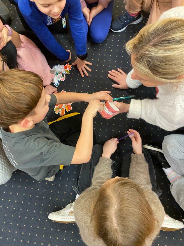 na zdjęciu widać grupę dzieci. na pierwszym planie chłopiec dotyka modelu jamy ustnej, który trzyma pani w blond włosach i białym sweterku.