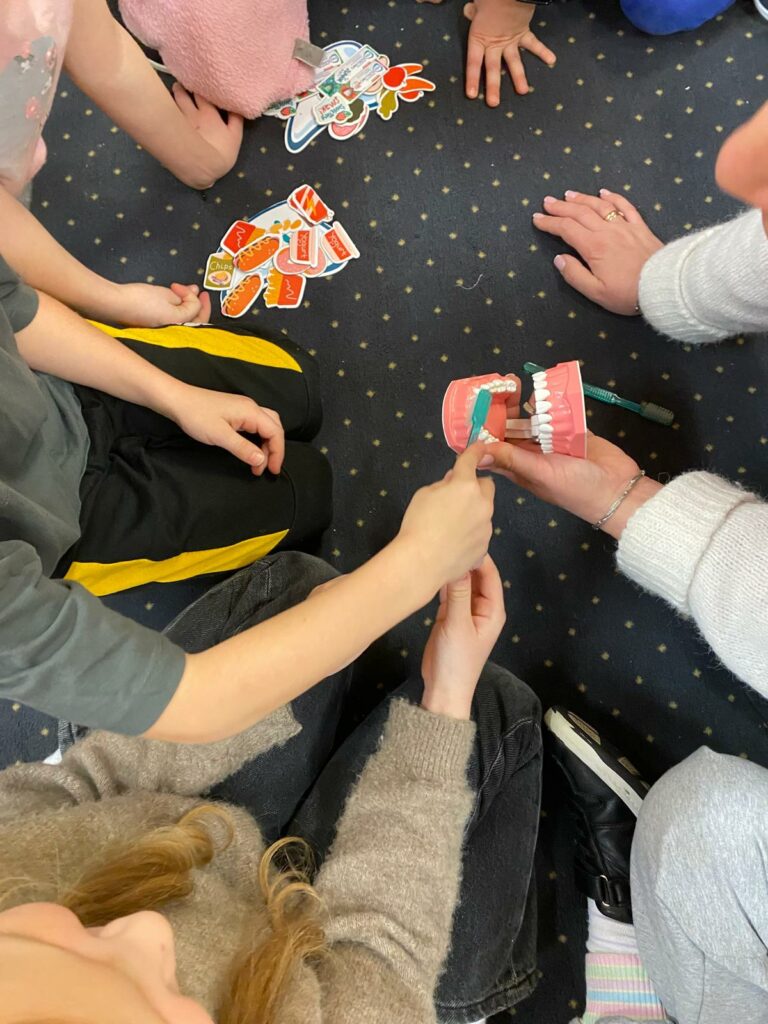 zdjęcie przedstawia wyciągniete ręce kilku dzieci. jedno dziecko dotyka szczoteczką modelu jamy ustnej. Na dywanie leżą obrazki z rzeczami zdrowymi dla zębów i niezdrowymi dla zębów.