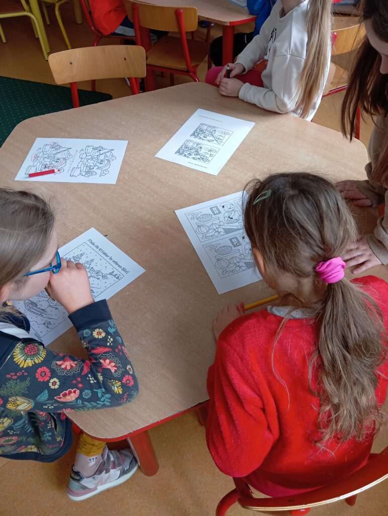 Na zdjęciu widać 4 dziewczynki siedzące przy stoliku. 1 dziewczynka ma długie brązowe włosy spięte różową gumką, ubrana jest w czerwony sweter.2 dziewczynka ma niebieskie okulary i kolorowy sweterek. 3 dziewczynka ma biały sweterek. 4 dziewczynka ma jest ubrana w brązową bluzę, ma ciemne włosy..Przed sobą mają kartki, na których mają znaleźć 10 różnic. Zabawa nazywa się „Bystre oko”