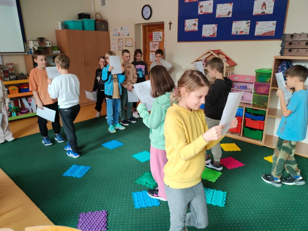 Na zdjęciu widać grupę dzieci w sali lekcyjnej, które trzymają kartki z tekstem. Zabawa nazywa się „Zaczytani”. Jej celem jest przejście przez labirynt podczas czytania. Na dywanie znajdują się kolorowe, sensoryczne maty, które dzieci mają ominąć a w tle widoczne są półki z zabawkami, materiały edukacyjne oraz tablica z uczniowskimi pracami.