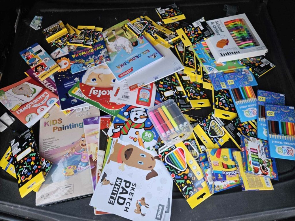 Na zdjęciu widoczny jest rozrzucony zestaw artykułów plastycznych i szkolnych. Znajdują się tam między innymi kolorowe kredki, flamastry, farby, zeszyty do rysowania, kolorowanki oraz różne zestawy papiernicze. Opakowania są kolorowe i przeznaczone głównie dla dzieci. Można zauważyć także plastelinę, bloki rysunkowe oraz różne pisaki.