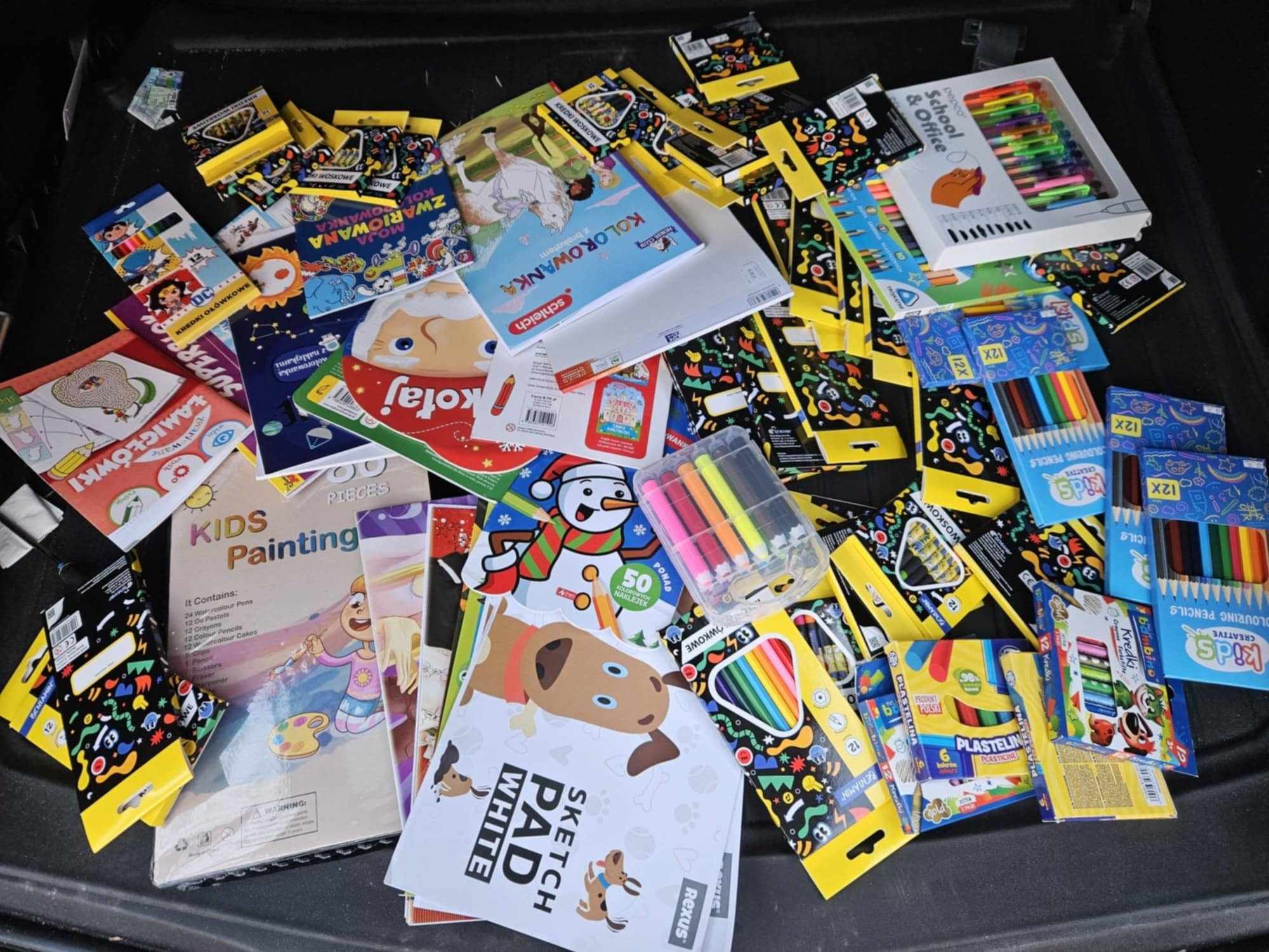 Na zdjęciu widoczny jest rozrzucony zestaw artykułów plastycznych i szkolnych. Znajdują się tam między innymi kolorowe kredki, flamastry, farby, zeszyty do rysowania, kolorowanki oraz różne zestawy papiernicze. Opakowania są kolorowe i przeznaczone głównie dla dzieci. Można zauważyć także plastelinę, bloki rysunkowe oraz różne pisaki.