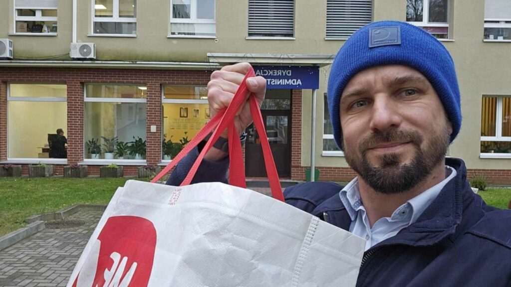 Na zdjęciu znajduje się mężczyzna z brodą, ubrany w niebieską czapkę i granatową kurtkę. Trzyma w ręku torbę z czerwono-białym W tle widać budynek z napisem „DYREKCJA ADMINISTRACYJNA”, co sugeruje, że znajduje sie przed budynkiem szpitala . Zdjęcie może przedstawiać moment dostarczania darów dla danej instytucji.