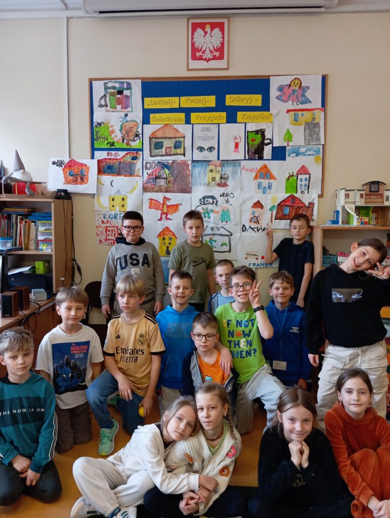 Grupa dzieci pozuje do zdjęcia w świetlicy szkolnej na tle tablicy z rysunkami przedstawiającymi „Moje bezpieczne miejsce”. 16 prac to dom, 1 praca KFC, 2 prace przedstawiają zajęcia taneczne, 1 praca przedstawia przebranie się za wróżkę. Na tablicy widnieje napis „Dotknij – Poczuj – Odkryj – Świetlicowa Przygoda Zmysłów”, Wszystkie są zadowolone, a niektóre przyjmują swobodne, radosne pozy, unoszą ręce do góry.