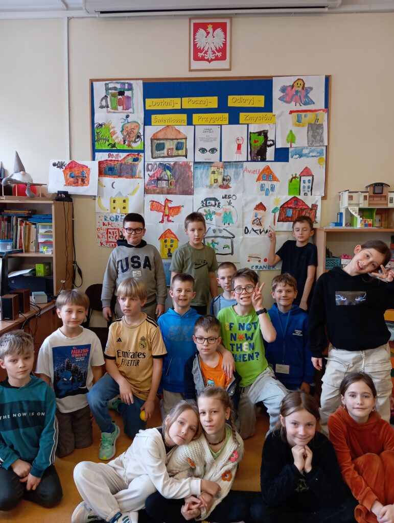 Grupa dzieci pozuje do zdjęcia w świetlicy szkolnej na tle tablicy z rysunkami przedstawiającymi „Moje bezpieczne miejsce”. 16 prac to dom, 1 praca KFC, 2 prace przedstawiają zajęcia taneczne, 1 praca przedstawia przebranie się za wróżkę. Na tablicy widnieje napis „Dotknij – Poczuj – Odkryj – Świetlicowa Przygoda Zmysłów”, Wszystkie są zadowolone, a niektóre przyjmują swobodne, radosne pozy, unoszą ręce do góry.