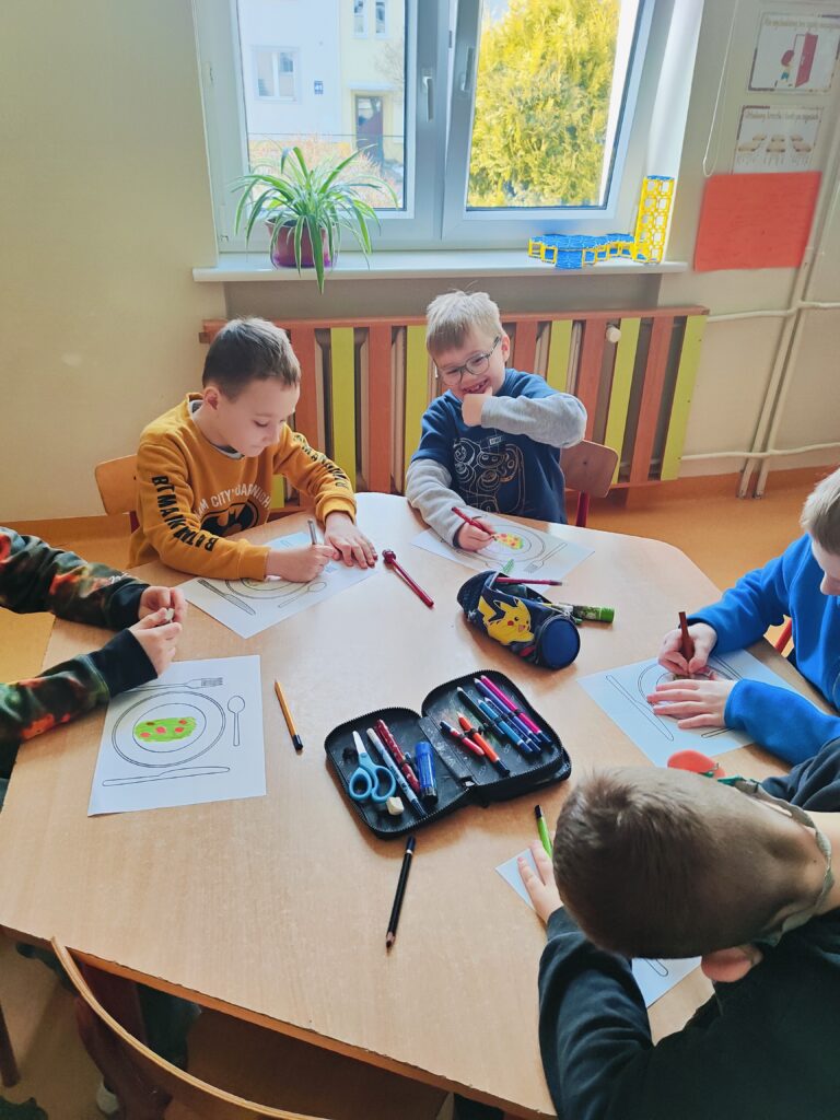 Na zdjęciu widzimy kilku chłopców siedzących przy sześciokątnym stole. Wykonują oni prace plastyczne. Na stole leżą kartki, na których przedstawione jest nakrycie stołu. Dwóch chłopców nachyla się nad kartką kolorując ją, jeden chłopiec w okularach uśmiecha się i dotyka dłonią brody . W tle widać okno, biały parapet i brązowo – zieloną obudowę do grzejnika.