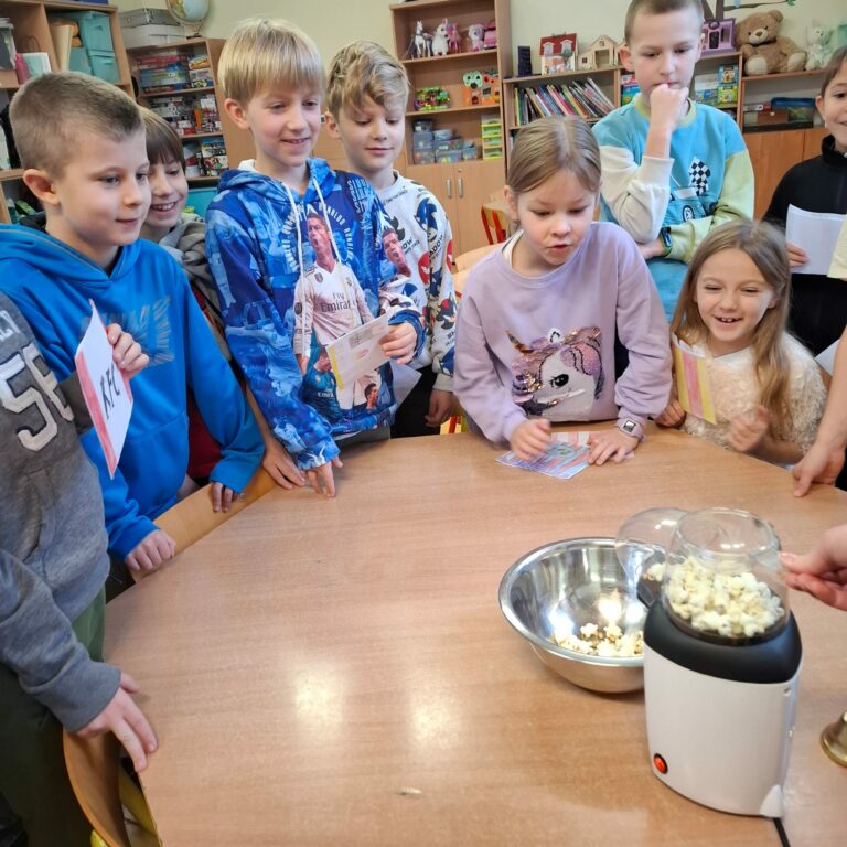 Na zdjęciu widać grupę dzieci zgromadzonych wokół stołu w sali lekcyjnej. Dzieci z zainteresowaniem obserwują działającą maszynę do robienia popcornu, z której świeży popcorn wpada do metalowej miski. Niektóre dzieci trzymają w rękach kolorowe kartki, a ich twarze wyrażają radość i ciekawość. W tle widać półki z zabawkami, książkami i grami, co sugeruje, że jest to sala szkolna lub świetlica.