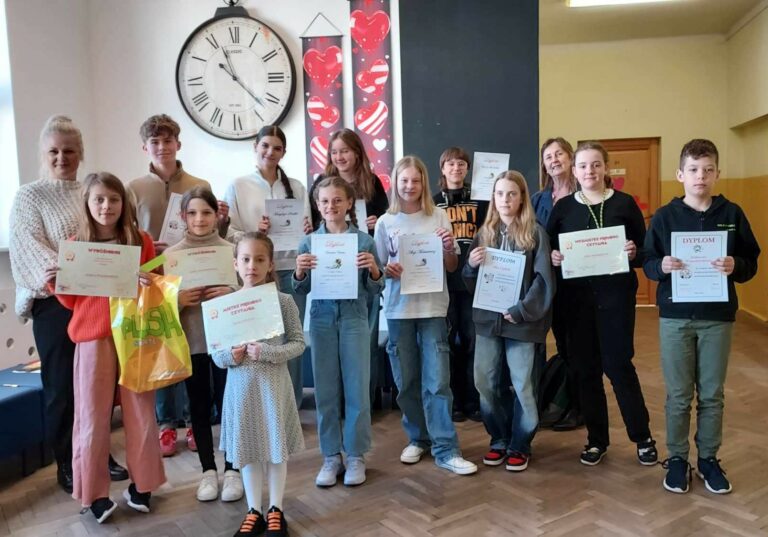 1. Na zdjęciu znajduje się grupa dzieci chłopców i dziewczęta wraz z dwiema dorosłymi kobietami. Dzieci trzymają w rękach dyplomy. Grupa stoi na szkolnym korytarzu – widać drewnianą podłogę, drzwi do sal lekcyjnych. W tle widoczny jest duży, okrągły zegar ścienny wskazujący godzinę 12:25, a obok niego dwie pionowe dekoracje z motywem czerwonych serc. Jedna z dziewczynek trzyma w dłoni oprócz dyplomu kolorową torbę. Grupa jest różnorodna wiekowo – od młodszych dzieci po nastolatków. Wszyscy na zdjęciu się uśmiechają. Strój uczestników jest codzienny, swobodny, ale niektóre osoby wybrały bardziej eleganckie elementy, jak białe koszule czy klasyczne swetry. Na stopach dominują sportowe buty.