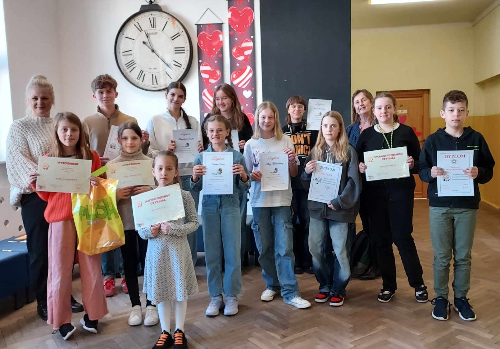 1. Na zdjęciu znajduje się grupa dzieci chłopców i dziewczęta wraz z dwiema dorosłymi kobietami. Dzieci trzymają w rękach dyplomy. Grupa stoi na szkolnym korytarzu – widać drewnianą podłogę, drzwi do sal lekcyjnych. W tle widoczny jest duży, okrągły zegar ścienny wskazujący godzinę 12:25, a obok niego dwie pionowe dekoracje z motywem czerwonych serc. Jedna z dziewczynek trzyma w dłoni oprócz dyplomu kolorową torbę. Grupa jest różnorodna wiekowo – od młodszych dzieci po nastolatków. Wszyscy na zdjęciu się uśmiechają. Strój uczestników jest codzienny, swobodny, ale niektóre osoby wybrały bardziej eleganckie elementy, jak białe koszule czy klasyczne swetry. Na stopach dominują sportowe buty.