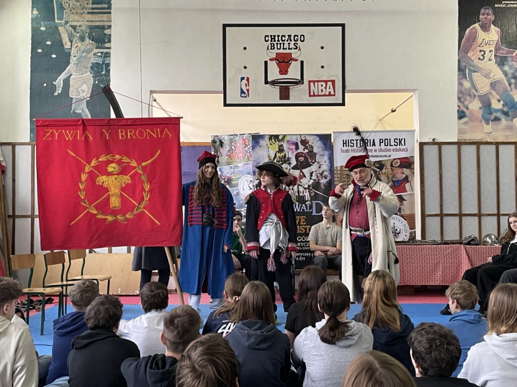 Na pierwszym planie stoi dwoje uczniów ubranych w kolorowe stroje kosyniera i Kościuszki. Jedno z nich trzyma czerwony sztandar. Obok stoi mężczyzna w stroju kosyniera.