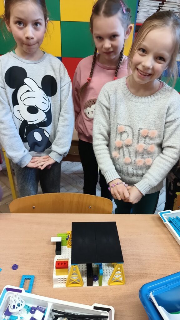 Trzy dziewczynki prezentują budowlę z klocków lego. Jest to czarno – biała konstrukcja przypominająca dom. Za dziećmi jest segment, na którym leżą książki.