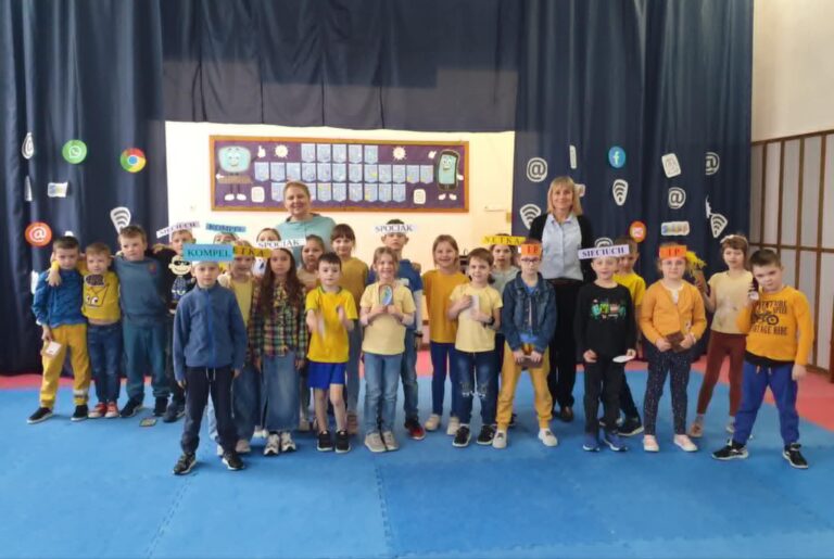 1. Na zdjęciu widzimy klasę 1a podczas apelu na temat bezpiecznego korzystania z Internetu. Uczniowie, ubrani głównie w żółte i niebieskie stroje, stoją na tle dekoracji z symbolami mediów społecznościowych oraz napisem "Dzień Bezpiecznego Internetu". Dzieci trzymają różne tabliczki z hasłami i nazwami związanymi z Internetem, a wraz z nimi stoją dwie nauczycielki, które wspierają edukację o cyberbezpieczeństwie.ciudzimy