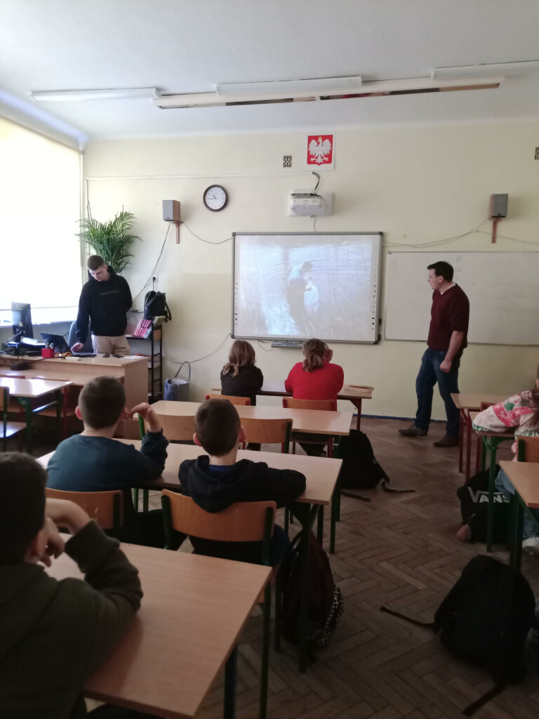 Na zdjęciu widać klasę siedzącą w ławkach oraz wyświetlaną na tablicy prezentacje