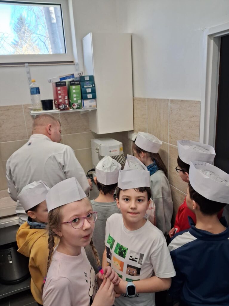 Na zdjęciu widać grupkę dzieci w pomieszczeniu kuchennym. Dwoje dzieci jest odwrócona twarza do obiektywu, reszta stoi tyłem. W głębi widać mężczyznę w stroju kucharza, obsługującego mikser. Nad nim wiszą na ścianie półki z kuchennym przyborami. Dzieci mają na głowie kucharskie czapki.