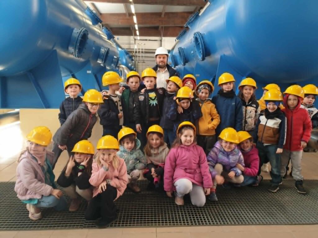 Na zdjęciu znajduje się grupa dzieci wraz z tatą Krzysia. wszyscy mają na głowach żółte kaski ochronne. Dzieci stoją i kucają w dużej hali otoczone ogromnymi, niebieskimi urządzeniami technicznymi. W tle widać elementy instalacji i oświetlenie sufitowe, co sugeruje, że znajdują się na wycieczce edukacyjnej w stacji uzdatniania wody. Atmosfera jest radosna i pełna ciekawości – dzieci uśmiechają się i z zainteresowaniem pozują do zdjęcia.