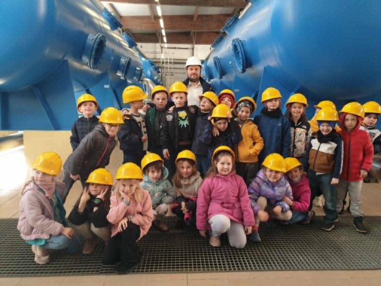 Na zdjęciu znajduje się grupa dzieci wraz z tatą Krzysia. wszyscy mają na głowach żółte kaski ochronne. Dzieci stoją i kucają w dużej hali otoczone ogromnymi, niebieskimi urządzeniami technicznymi. W tle widać elementy instalacji i oświetlenie sufitowe, co sugeruje, że znajdują się na wycieczce edukacyjnej w stacji uzdatniania wody. Atmosfera jest radosna i pełna ciekawości – dzieci uśmiechają się i z zainteresowaniem pozują do zdjęcia.