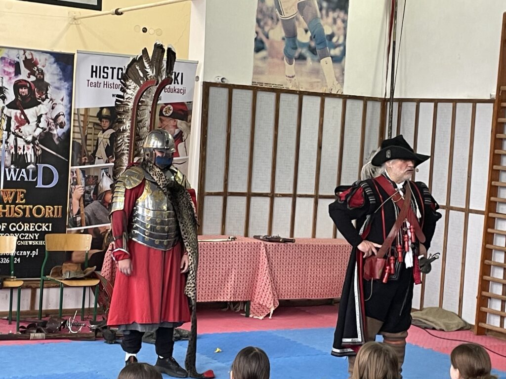 Na środku stoi chłopiec w stroju husarza. Ma na sobie zbroję i skrzydła.