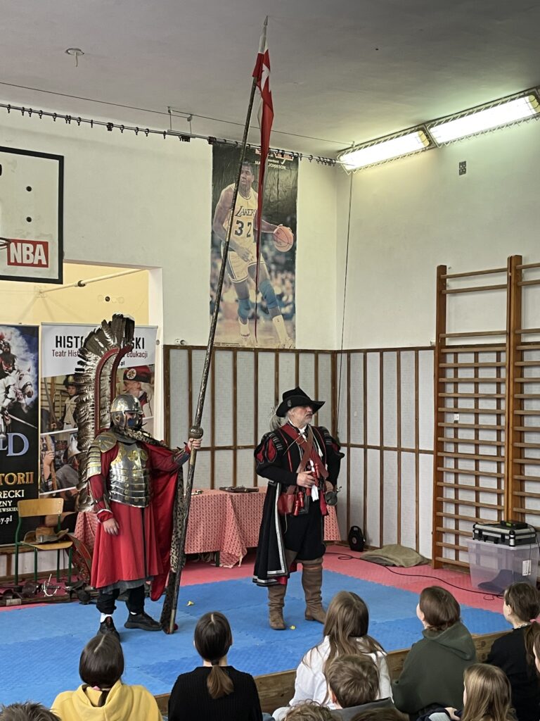 Na środku stoi chłopiec w stroju husarza. Ma na sobie zbroję i skrzydła a w ręku trzyma pionowo kopię husarską.