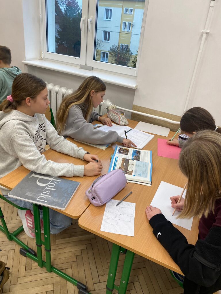 Klasa szkolna, okno, kaloryfer, ławki, czworo dzieci, dziewczynki, 12 lat ubrane w szare i czarne bluzy, na ławkach zeszyty, książki, piórniki