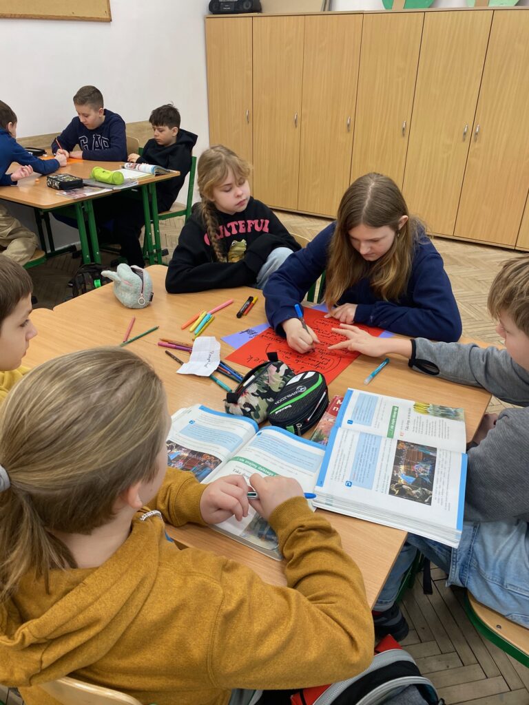 Klasa szkolna, szafy, ławki, ośmioro dzieci, dziewczynki i chłopcy 12 lat ubrane w szare, czarne, żółte, granatowe bluzy, na ławkach zeszyty, książki, piórniki.