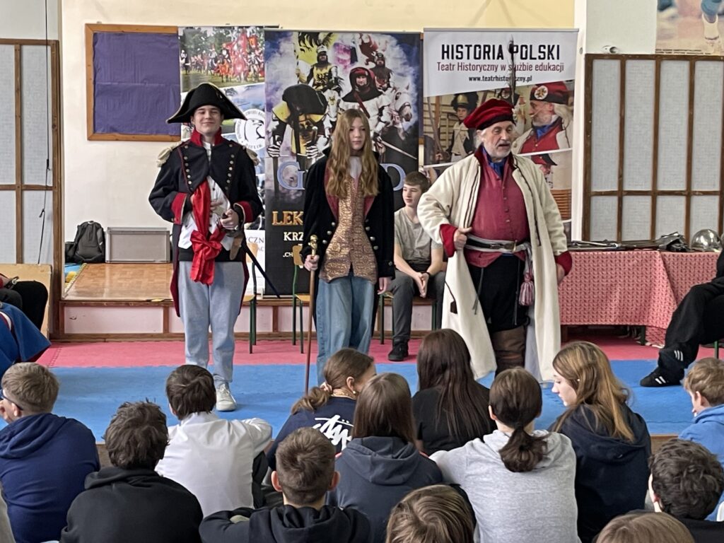 N środku stoi chłopiec przebrany za cesarza napoleona oraz dziewczyna w stroju Józefa Wybickiego. Obok stoi mężczyzna , który wyjaśnia dzieciom treści pokazu historycznego.