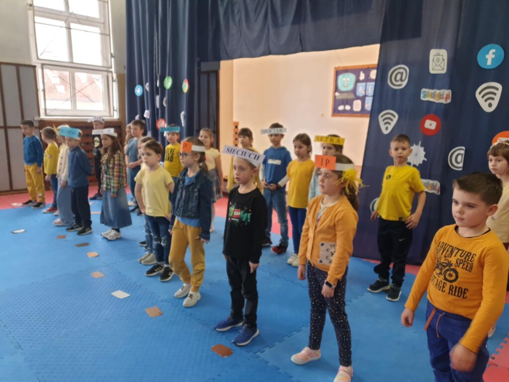 Na zdjęciu widzimy występ uczniów pierwszej klasy na temat bezpiecznego korzystania z Internetu. Dzieci ubrane są w żółte i niebieskie ubrania, a na głowach mają opaski z napisami nawiązującymi do sieci i technologii, co sugeruje edukacyjny charakter przedstawienia. Dekoracja z ikonami mediów społecznościowych i internetu podkreśla tematykę wydarzenia, które prawdopodobnie ma na celu nauczenie dzieci zasad cyberbezpieczeństwa w przystępnej formie.