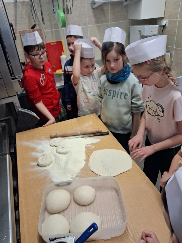 W kuchni znajduje się grupa dzieci. Mają na głowach papierowe czapki kucharskie. Na drewnianych blacie leży rozwałkowane ciasto oraz kilka kulek przygotowanych do rozwałkowania. jedna osoba rozwałkowała swoje ciasto,reszta osób patrzy. W tle widoczne urządzenia kuchenne, takie jak sztućce, patelnie oraz pojemniki.