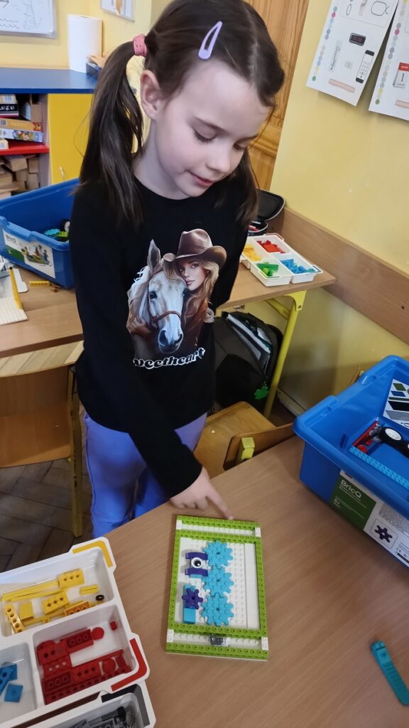 Dziewczynka ubrana w czarna bluzkę prezentuje budowlę z klocków lego. Jest to niebiesko – zielona konstrukcja przypominająca ogród. Za dzieckiem są ławki szkolne oraz półka z książkami.