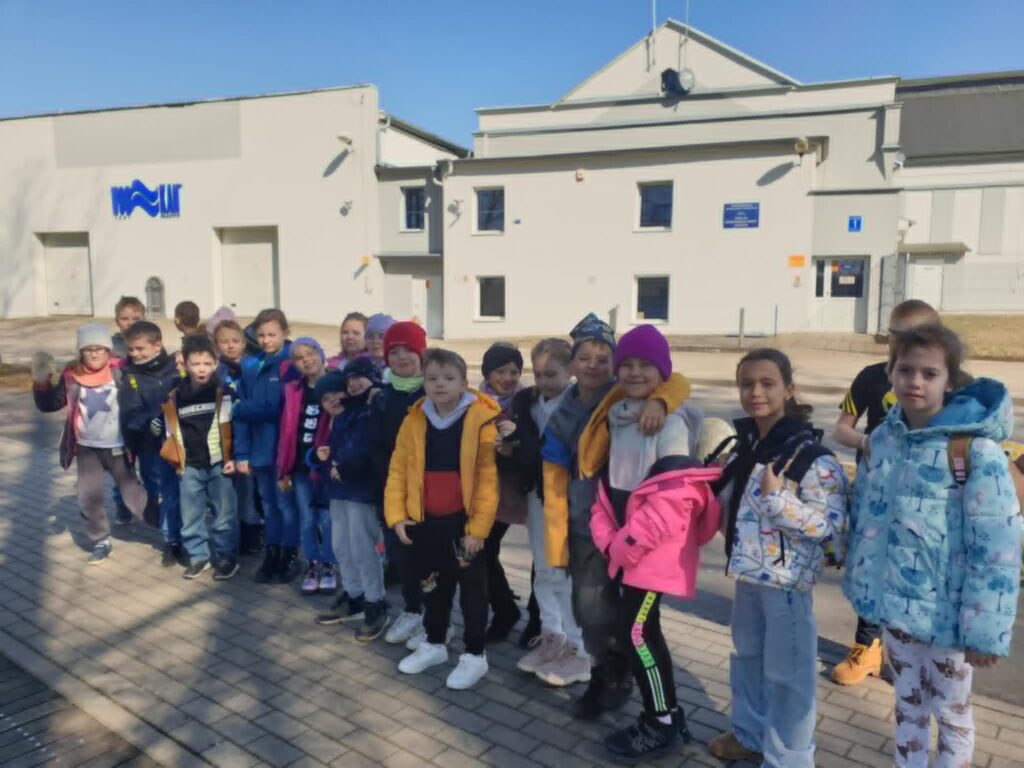 Na zdjęciu znajduje się grupa dzieci stojących w szeregu na chodniku, przed budynkiem o jasnej elewacji. Dzieci są ubrane w kolorowe kurtki i czapki, co sugeruje chłodną, ale słoneczną pogodę. W tle widać budynek z napisem "Wod-Kan" oraz niebieską tabliczką informacyjną. Wszystkie dzieci wyglądają na uśmiechnięte i zadowolone, co może wskazywać na wycieczkę lub wyjście szkolne.