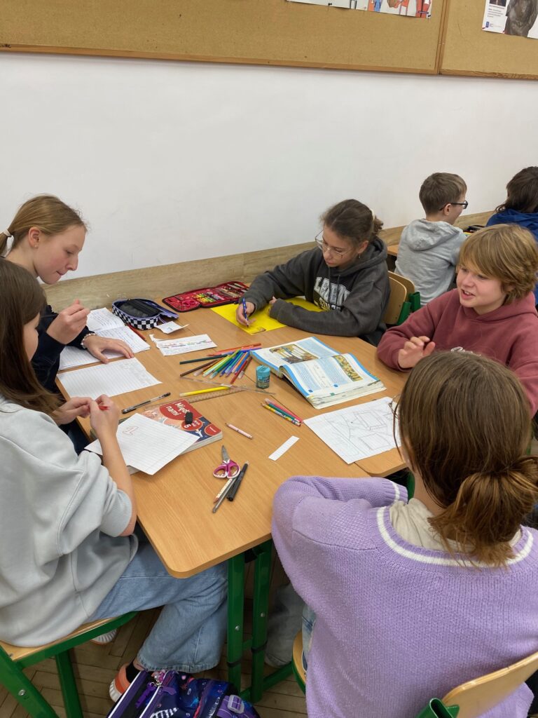 Klasa szkolna, ławki, tablice korkowe, siedmioro dzieci, dziewczynki i chłopcy 12 lat ubrane w szare, czarne, fioletowe, granatowe, bordowe bluzy, na ławkach zeszyty, książki, piórniki.