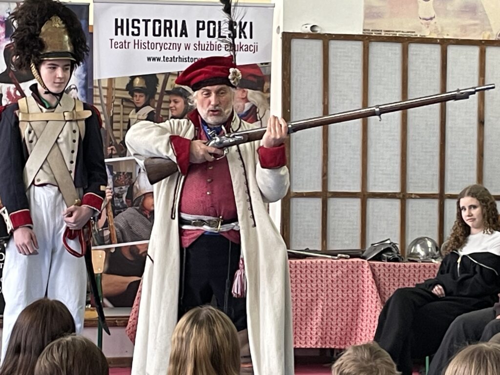 Na środku stoi chłopiec w mundurze z czasów Księstwa Warszawskiego, obok mężczyzna pokazuje strzelanie z ówczesnej broni.