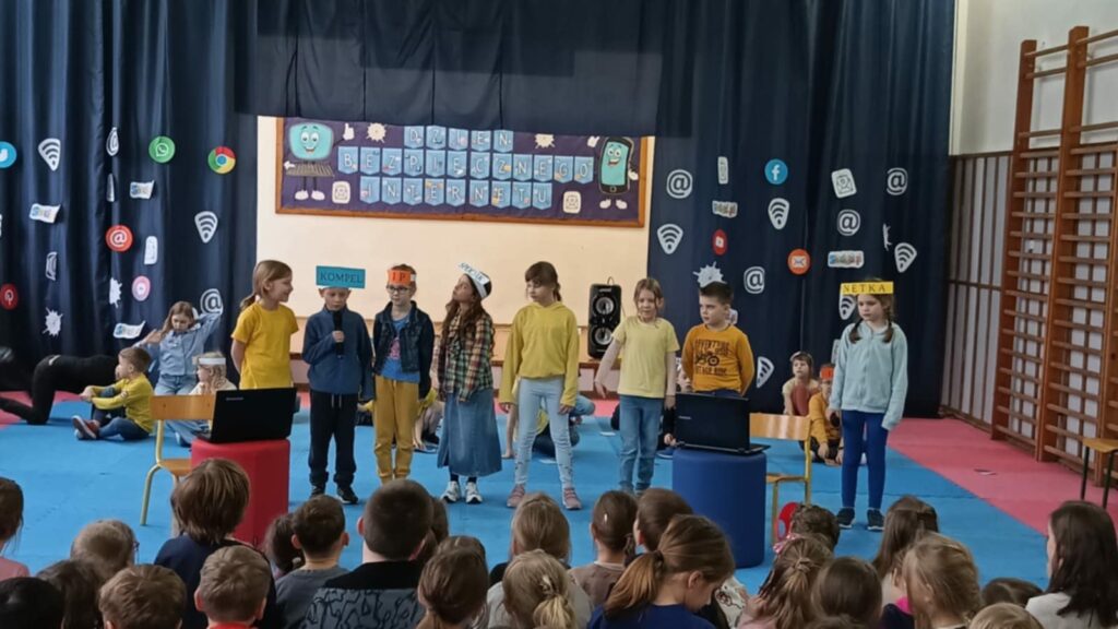 Zdjęcie przedstawia apel szkolny związany z obchodami Dnia Bezpiecznego Internetu. Grupa dzieci na scenie bierze udział w przedstawieniu, mając na głowach opaski z napisami nawiązującymi do internetu, a dekoracje zawierają symbole mediów społecznościowych i komunikacji online. Przed nimi widoczna jest publiczność składająca się z uczniów, którzy oglądają występ.