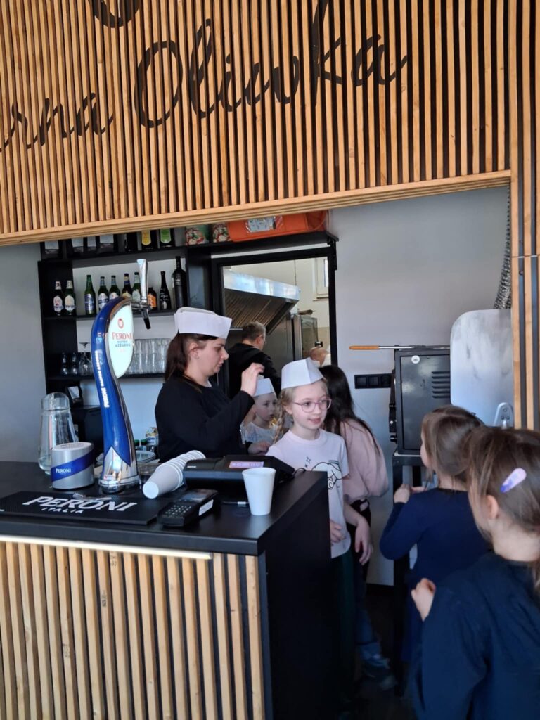 Na zdjęciu widać drewniany kontuar i fragment ściany z logo restauracji. Za ladą stoi kobieta i kilkoro dzieci, które wychodza z kuchni. Dwoje z nich ma na głowach ma kucharskie czapki.