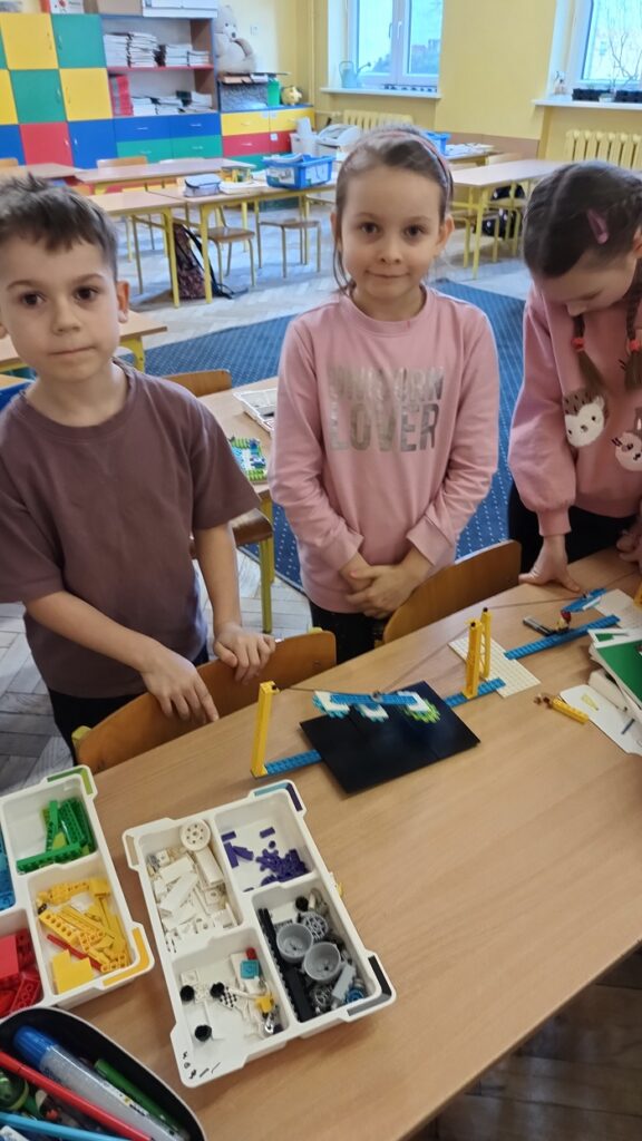 Troje dzieci - dwie dziewczynki i jeden chłopiec prezentują budowlę z klocków lego. Jest to czarno – biała konstrukcja przypominająca skocznię narciarską. Za dziećmi jest segment, na którym leżą książki oraz ławki i krzesła.