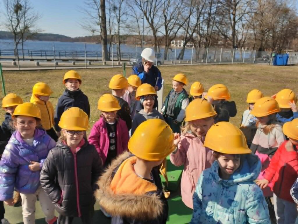 Na zdjęciu znajduje się grupa dzieci noszących żółte kaski ochronne, stojących na świeżym powietrzu. W tle widać jezioro, ogrodzenie oraz drzewa bez liści, co pokazuje, że to wczesną wiosnę. Dzieci są ubrane w ciepłe kurtki, a wśród nich znajduje się osoba dorosła w białym kasku, - przewodnik po obiekcie.