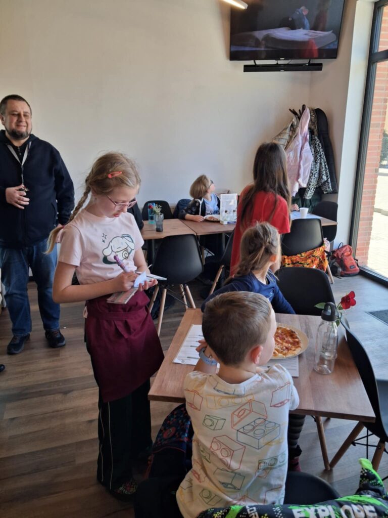 W sali restauracyjnej, przy stolikach siedzą dzieci. Na stoliku leży pizza. Obok stolika stoi dziewczynka w fartuszku i czapce kucharskiej. Pisze w notesie i zbiera zamówienia od dzieci. W głębi stoi mężczyzna.