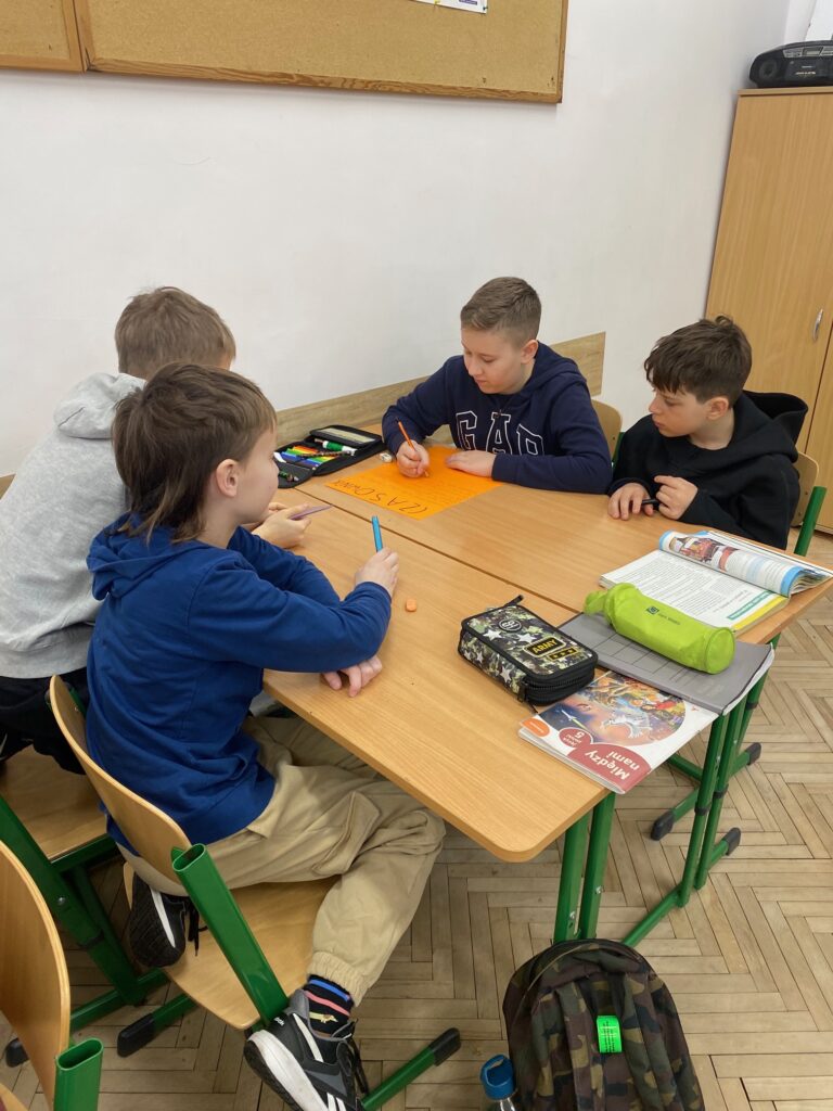 Klasa szkolna, szafy, tablice korkowe, ławki, ośmioro dzieci, chłopcy 12 lat ubrani w szare, czarne, granatowe bluzy, na ławkach zeszyty, książki, piórniki.