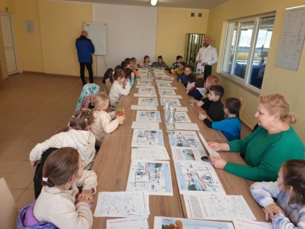 Na zdjęciu widać grupę dzieci siedzących przy długim stole w sali edukacyjnej. Na stole leżą plansze edukacyjne, a dzieci słuchają prezentacji prowadzonych przez dwóch dorosłych mężczyzn. Jedna z kobiet siedzi razem z dziećmi i również przegląda materiały edukacyjne.