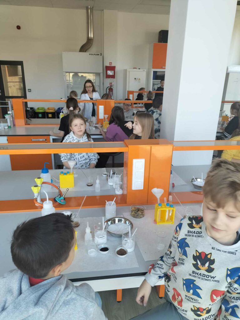 Dzieci siedzą przy pulpitach roboczych, na których zgromadzono różne substancje i przybory laboratoryjne.