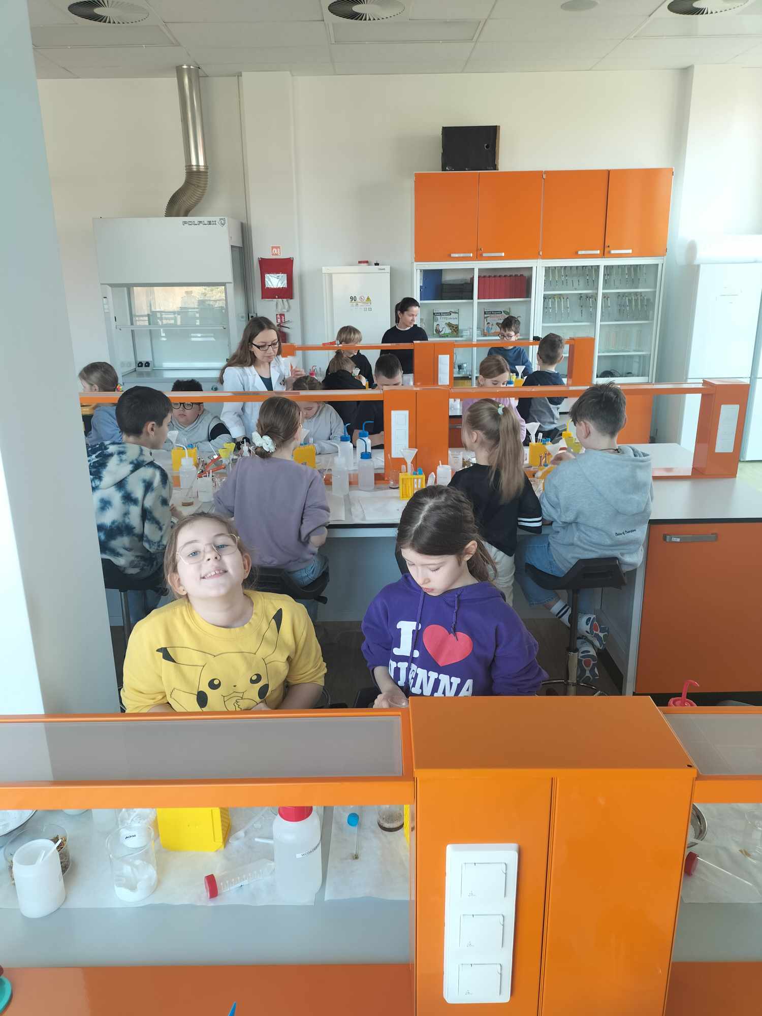 Dzieci siedzą przy pulpitach roboczych, na których zgromadzono różne substancje i przybory laboratoryjne.