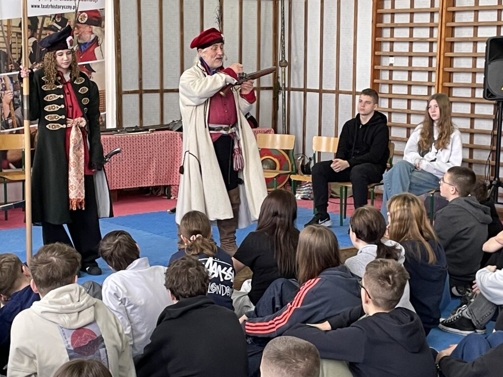 Na środku stoi dziewczyna przebrana za powstańca styczniowego. Obok mężczyzna pokazuje broń.