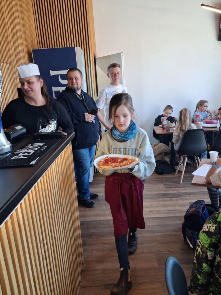 W sali restauracyjnej na pierwszym planie widać dziewczynkę w fartuszku. Niesie pizzę na talerzu. Po prawej stronie widać dzieci, siedzące pray stolikach. Po lewej widać drewniany kontuar, a za nim stoi kobieta w czapce kucharskiej. Za dziewczynką, w głębi widać dwóch mężczyzn.