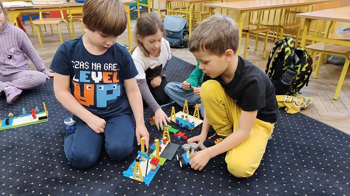 Dwóch chłopców i dziewczynka siedzą na granatowym dywanie. Dzieci budują konstrukcję z klocków lego. Za nimi są ławki i krzesła.