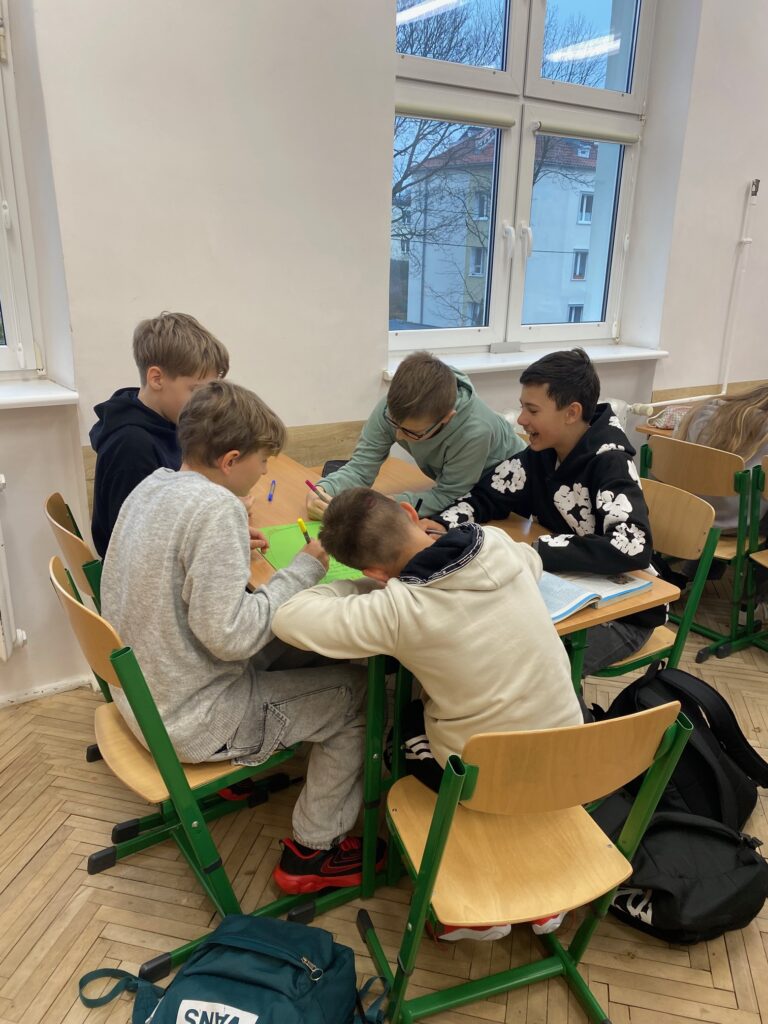 Klasa szkolna, okno, ławki, pięcioro dzieci, chłopcy 12 lat ubrani w szare, czarne, zielone, granatowe bluzy, na ławkach zeszyty, książki, piórniki.