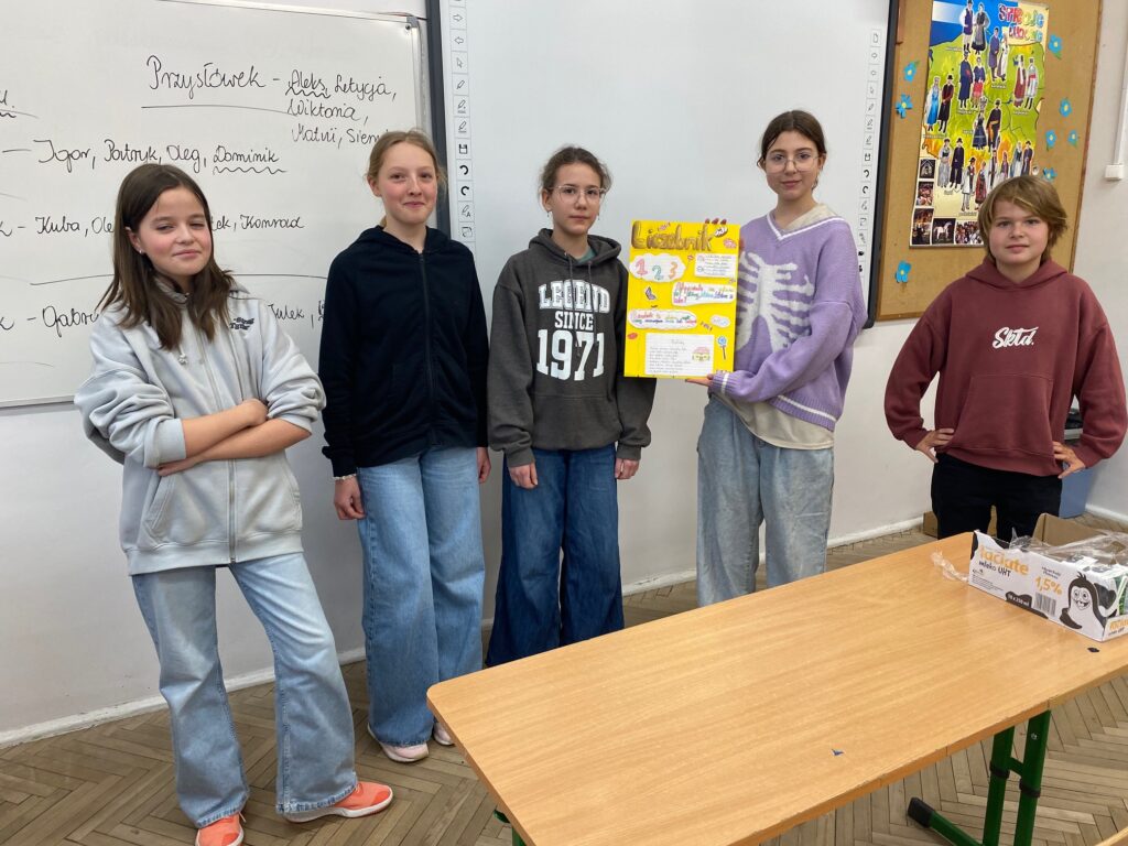Klasa szkolna, tablica, ławki, pięcioro dzieci, dziewczynki i chłopcy 12 lat ubrani w szare, czarne, fioletowe, bordowe bluzy, trzymają plakat, na ławce mleko w kartonikach.